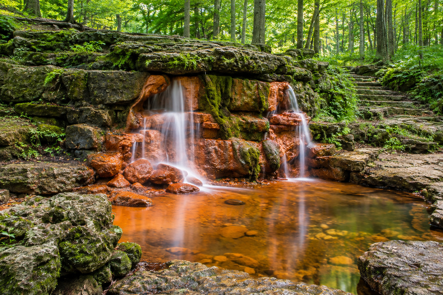 A mossy forest waterfall over rust-colored rock into a still pool, with stone steps through the trees.